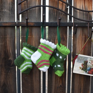 May include: Three small, knitted Christmas stockings in shades of green, white, and pink hang from a decorative metal hook. Each stocking has a unique pattern, including stripes and polka dots, and is suspended by a green ribbon.
