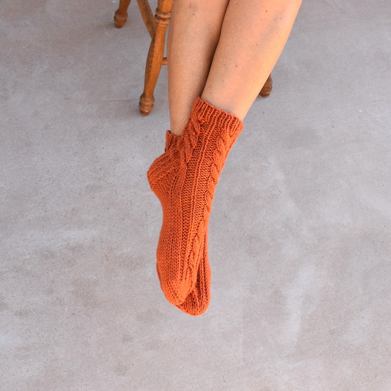 May include: A pair of orange knitted socks with a cable knit design. The socks are a warm, earthy tone and appear to be handmade. The socks are visible from the ankle down, with a wooden chair and legs in the background.