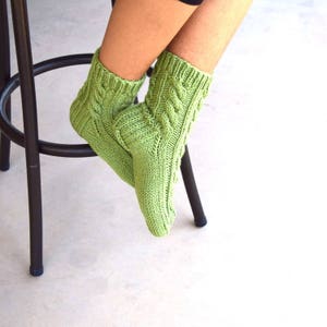 May include: A pair of green knitted socks with a cable knit pattern. The socks are worn on a person's feet and are sitting on a black metal stool.
