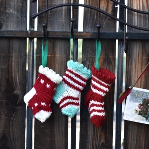 May include: Three knitted Christmas stockings in red, white, and teal, hanging from a black metal hook. The stockings have various patterns, including stripes and hearts, and are suspended by green ribbons. A festive holiday decoration.
