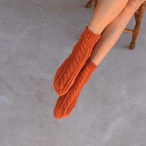 May include: A pair of orange cable-knit socks. The socks are a warm, burnt orange color and feature a detailed cable knit pattern. The socks are shown on a person's legs, with the feet resting on a gray surface.