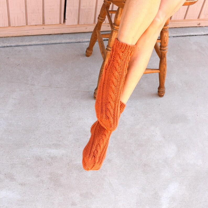 May include: A pair of orange cable-knit socks. The socks are a warm, burnt orange color and feature a classic cable knit design. The socks are knee-high and appear to be handmade. The background includes a wooden stool and a pink wall.