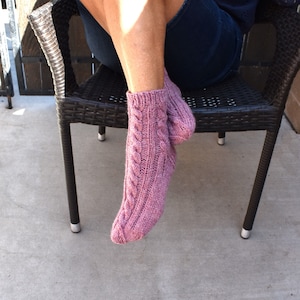May include: A pair of pink knitted socks with a cable knit pattern. The socks are worn on a person's feet.