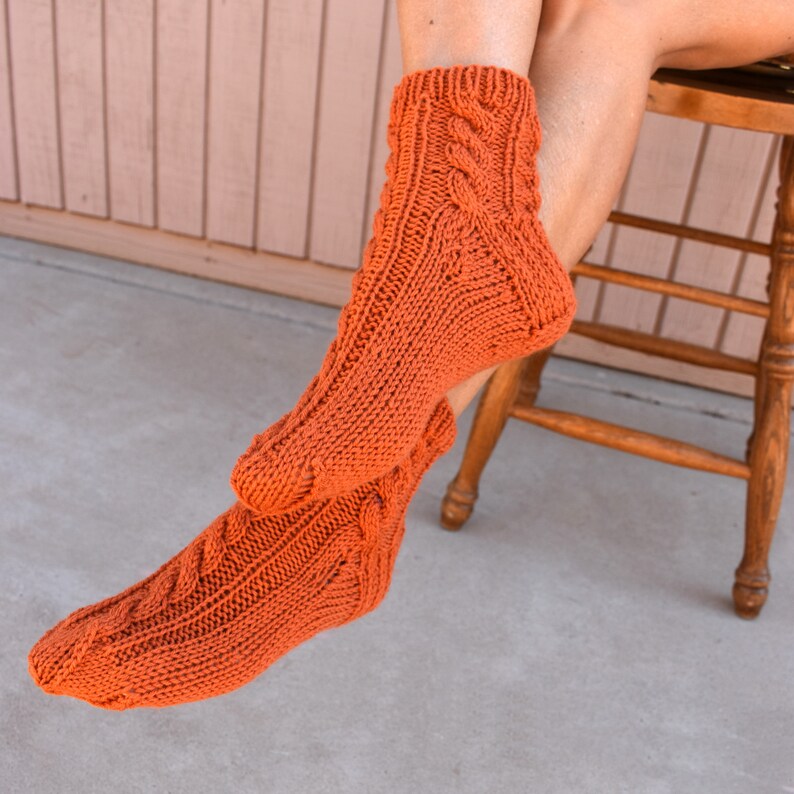 May include: A pair of orange knitted socks with a cable knit design. The socks are a warm, burnt orange color and appear to be handmade. The socks are worn on a person's feet, with the legs and a wooden chair visible in the background.