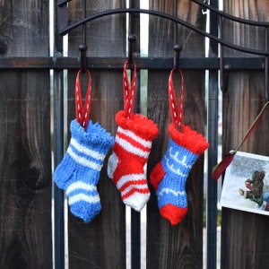 May include: Three miniature knitted Christmas stockings in blue, red, and white hang from a black metal hook. Each stocking is adorned with a red ribbon for hanging. The stockings feature various patterns, including stripes and snowflakes.