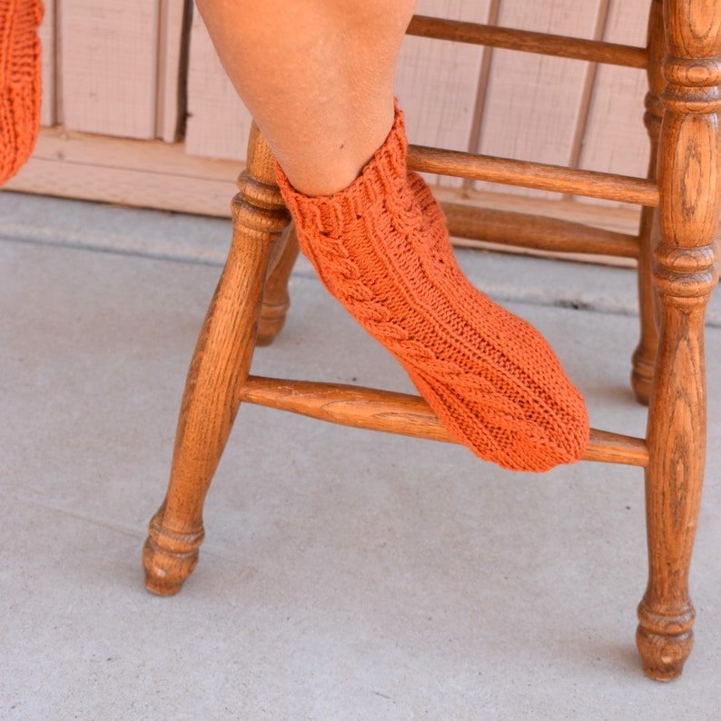 May include: Close-up of an orange knitted sock with a cable knit design. The sock is worn on a foot resting on a wooden chair. The sock's color is a vibrant orange.