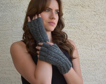 May include: Gray fingerless gloves with a cable knit design. The gloves are made of a soft, knitted material. The gloves cover the hand up to the knuckles, leaving the fingers free. The gloves are a neutral color and can be worn with many outfits.