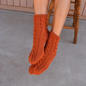 May include: A pair of orange cable-knit socks. The socks are a warm, burnt orange color and feature a detailed cable knit pattern. The socks are thick and appear to be handmade, perfect for cold weather.