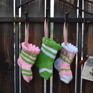 May include: Three miniature knitted Christmas stockings in pink, green, and white hang from a dark metal hook. The stockings have striped and scalloped designs, with red and white striped ribbon for hanging. The background is a wooden fence.