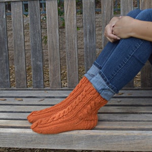 May include: A pair of orange knitted socks with a cable knit pattern. The socks are worn on a person's feet and are resting on a wooden bench.