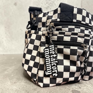 May include: Black and white checkered crossbody bag with multiple zippered compartments. The bag features a black strap and a keychain that reads "metalcore mommy" in white lettering. The bag is made of a corduroy-like material.
