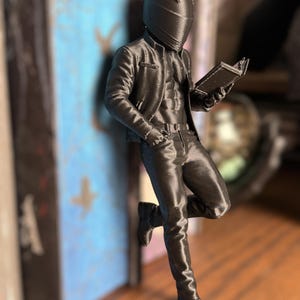 Könnte beinhalten: Eine schwarze Figur einer Person mit Motorradhelm und Jacke, die ein Buch liest und sich an eine Wand lehnt. Die Figur hat detaillierte Muskeldefinitionen und realistische Kleidung.