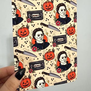 May include: A rectangular card featuring a repeating pattern on a cream background. The design includes illustrations of a masked figure, orange jack-o'-lanterns, silver knives, and black VHS tapes labeled HALLOWEEN, accented with small red flowers.