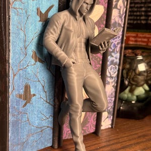 Pu&ograve; includere: Figurina grigia stampata in 3D di un uomo con felpa con cappuccio e jeans, appoggiato a un libro. L'uomo tiene in mano un libro e sembra leggere. Il dorso del libro ha uno sfondo blu con un disegno di albero e uccello.