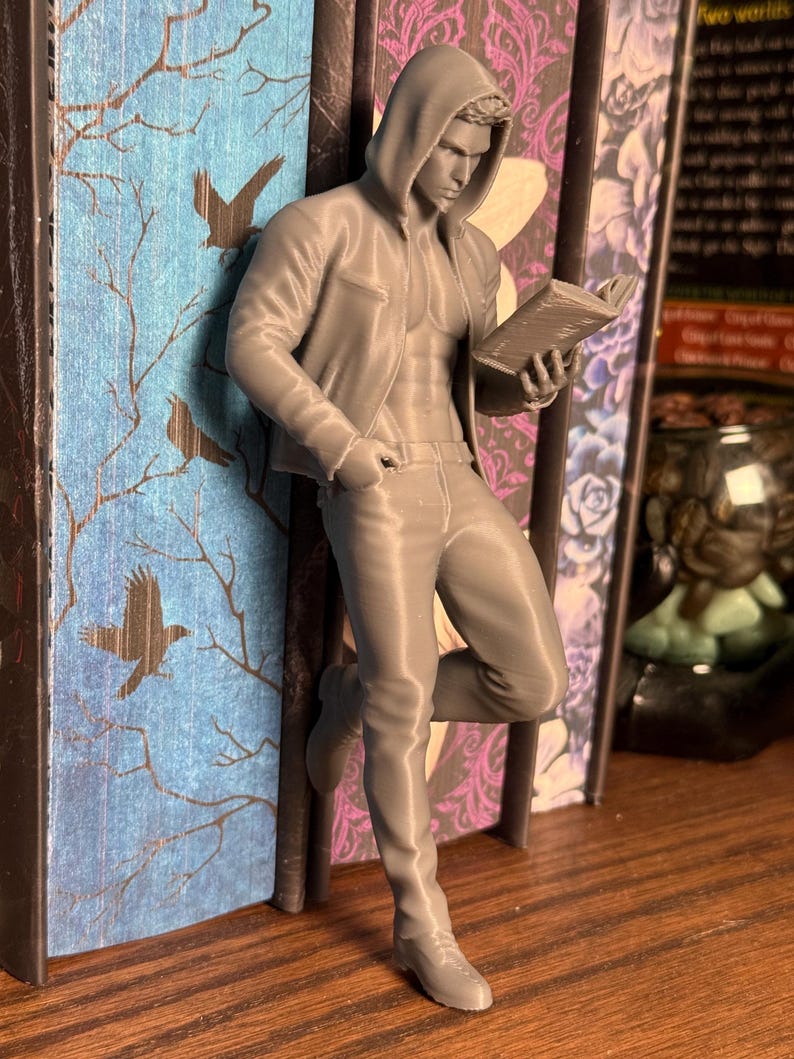 Pu&ograve; includere: Figurina grigia stampata in 3D di un uomo con felpa con cappuccio e jeans, appoggiato a un libro. L'uomo tiene in mano un libro e sembra leggere. Il dorso del libro ha uno sfondo blu con un disegno di albero e uccello.