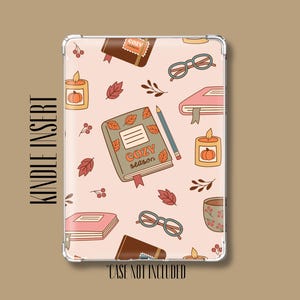 May include: A rectangular Kindle insert featuring a light pink background with a repeating pattern of books, candles with pumpkins, eyeglasses, pencils, and autumn leaves. The central book displays the text "COZY season" in orange lettering.