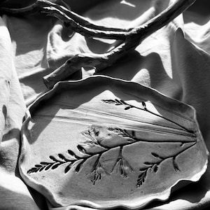 May include: A black and white image of a ceramic tray with a raised design of a plant. The tray is on a white linen fabric.