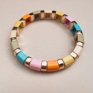 May include: A colourful bracelet made of square beads in shades of yellow, orange, pink, blue, green, and cream. The beads are separated by gold-toned spacers.