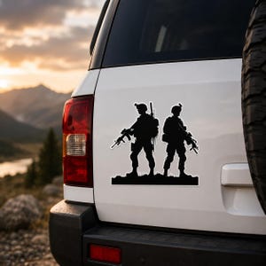 以下が含まれることがあります： 2人の兵士の黒いシルエットステッカー。それぞれがライフルを持って並んで立っています。ヘルメットをかぶり、バックパックを背負っています。ステッカーは白い車の後部に貼られており、背景には山と夕焼けの風景が広がっています。