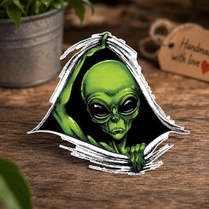 Può includere: Un alieno verde sbircia attraverso uno sfondo nero strappato. L'alieno ha grandi occhi neri e tiene i bordi dello strappo. L'immagine è un adesivo o una decalcomania, con un effetto carta strappata.