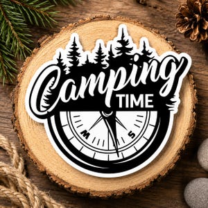 Op de afbeelding: Een zwart-witte sticker met de woorden "Camping Time" boven een kompasontwerp. De sticker heeft een silhouet van dennenbomen en is geplaatst tegen een houten achtergrond, met dennennaalden en een dennenappel zichtbaar.
