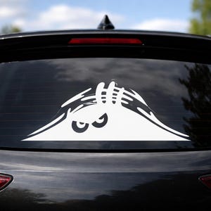 Op de afbeelding: Witte vinyl auto sticker met een monster ontwerp. De sticker toont een paar ogen die over een bergvorm met klauwhanden kijken. De sticker is aangebracht op de achterruit van een zwarte auto.