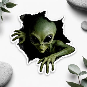 Puede incluir: Una pegatina troquelada que muestra a un alienígena verde con grandes ojos negros emergiendo de un agujero oscuro y dentado. El alienígena tiene una cabeza lisa y texturizada y dedos largos y delgados que se extienden desde el vacío.