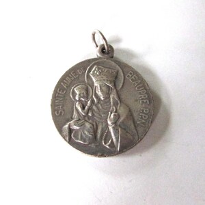May include: Silver-toned metal pendant featuring a depiction of Sainte Anne de Beaupre, a Catholic saint, holding a child. The pendant has the text "Sainte Anne de Beaupre" inscribed around the edge.