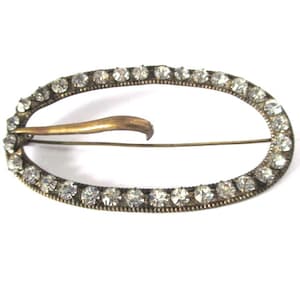May include: A gold-toned oval brooch with a row of clear rhinestones around the edge.
