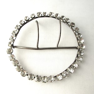May include: A silver-toned metal belt buckle with a circular frame adorned with clear rhinestones.