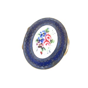 May include: A small, oval-shaped, blue enamel trinket box with a white enamel interior. The box is decorated with a hand-painted floral design in shades of red, blue, and green.
