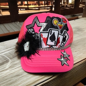 May include: A bright pink trucker hat with a mesh back and a curved brim. The hat is decorated with playing cards, dice, a star, and a black feather accent. Silver star and chain details add sparkle.