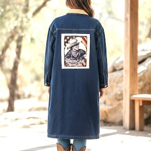 May include: A long, dark blue denim jacket with a cream-colored shearling collar. The back of the jacket features a rectangular patch with an illustration of a cowgirl and the words "AMERICAN COWGIRL". The person is wearing brown cowboy boots.