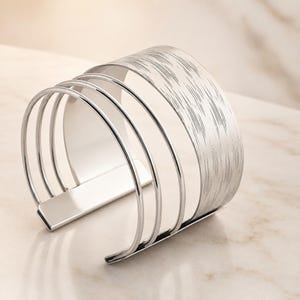 May include: A silver-toned cuff bracelet featuring a multi-strand design. The piece consists of three thin, smooth circular bands connected to a wider, flat band that displays a textured, brushed finish across its curved surface.