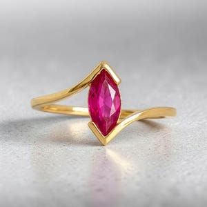 May include: A gold ring featuring a single marquise-cut vibrant pink gemstone set in a V-shaped prong setting. The band is smooth and polished, curving gently to meet the central stone, creating a sleek and continuous silhouette for the jewellery piece.