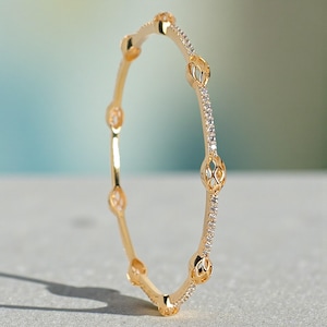 Può includere: Un braccialetto rigido dorato con un design dettagliato. Il braccialetto presenta sezioni traforate e incastonate con gemme alternate. Le gemme sono piccole e chiare, aggiungendo brillantezza. Il braccialetto è curvo.