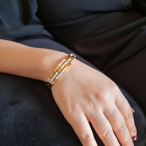 May include: Gold-toned bracelet with a layered design, featuring sparkling white gemstones. The bracelet is worn on a wrist, showcasing its intricate details and elegant style. The jewelry piece is a statement accessory.