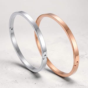 Może przedstawiać: Dwie bransoletki typu bangle na zawiasach, jedna w kolorze polerowanego srebra, a druga w kolorze polerowanego różowego złota. Każda bransoletka ma gładką, płaską powierzchnię i bezpieczne zapięcie, zaprojektowane jako prosta, okrągła obręcz.