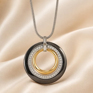 May include: A pendant necklace featuring a large black circular ring, a smaller gold-toned inner ring, and a band of clear crystals set between them. The pendant hangs from a silver-toned snake chain, connected by a rectangular crystal bail.