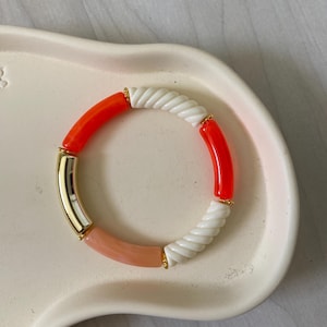 Puede incluir: Una pulsera colorida con cuentas naranjas, blancas, rosas y doradas. La pulsera presenta una combinación de cuentas lisas y texturizadas, creando un accesorio elegante. Circunferencia aproximada de 19 cm.