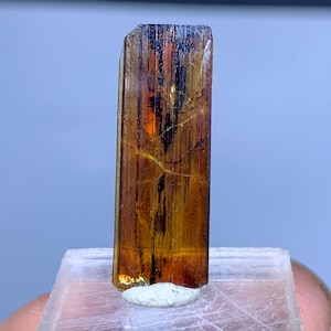 Puede incluir: Un espécimen mineral rectangular y translúcido con un rico tono marrón ámbar, acentuado por inclusiones lineales oscuras. El cristal está montado sobre una base blanca, mostrando su textura natural y sin pulir. El color del mineral pasa de marrón oscuro a un tono dorado más claro.