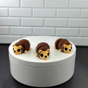May include: Three small, brown hedgehog figurines with yellow faces and paws are displayed on a white, circular platform. The hedgehogs have a spiky texture on their backs and are set against a white brick wall background.