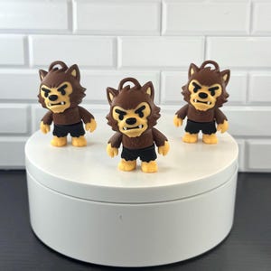 May include: Three small werewolf figurines with brown fur, pointed ears, and black shorts. Each character has a tan face, black eyebrows, and visible white fangs. The figures are standing upright and feature a small loop at the top of their heads.