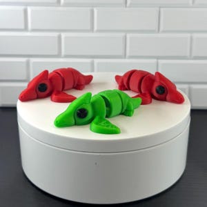 May include: Three articulated animal figurines, two in red and one in green, each featuring a segmented body, a smooth surface, and prominent black circular eyes. The figures are positioned on a white circular platform with a flat top and sides.
