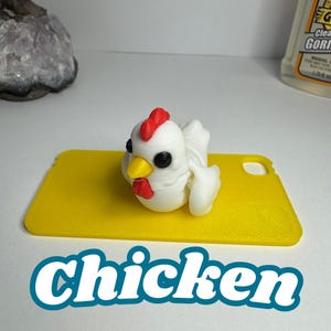 May include: A white chicken figurine with a red comb and yellow beak sits on a yellow rectangular base. The word "Chicken" is written in a teal font below the base. The figurine has black eyes and white wings.
