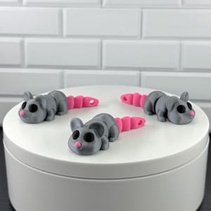 May include: Three small, articulated mouse figurines made of gray plastic with pink noses and segmented pink tails. Each mouse features large, round black eyes and a smooth, matte finish, arranged in a triangular formation on a white surface.