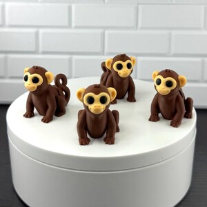 May include: Three small monkey figurines made of brown and cream-colored material. Each monkey has large black eyes, a light-colored face, and a curled tail. The figures are positioned in a group on a smooth, white, circular surface.