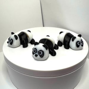 May include: Four small panda figurines. The pandas are black and white, with black ears, eyes, and limbs. The pandas are lying down on a white surface. The figurines are likely made of plastic or resin.