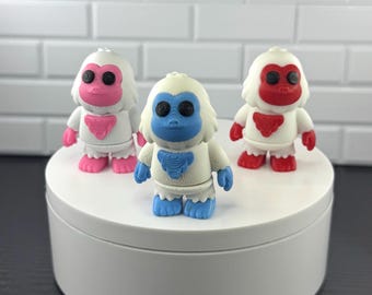 May include: Three small white yeti figurines with black circular eyes. The left figure has pink facial and chest accents, the center figure has blue accents, and the right figure has red accents. Each figure features a textured, molded design.