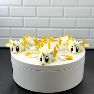 May include: A white cylindrical container topped with three small, white, dragon-like figurines. Each figurine features large black eyes, a single gold horn on its forehead, and gold-tipped wings and spines, arranged symmetrically on the lid.